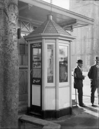 A black and white photograph from the early 20th century of two men standing outside a public phone box in Christchurch