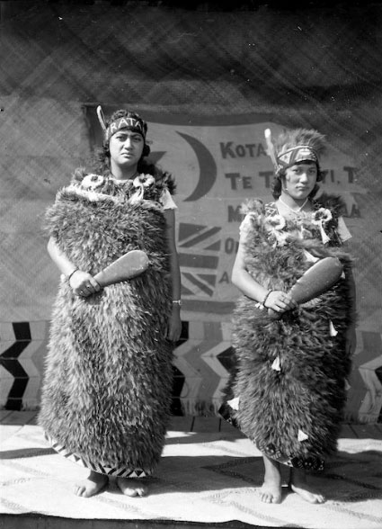 A black and white photo from 1924 of two Māori women in kiwi-feather kahu and holding mere.