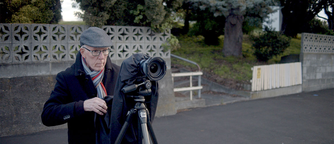 An older man, dressed against the cold on a quiet street, prepares a camera on a tripod.