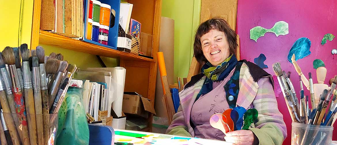 A woman sits surrounded by paintbrushes, paint and other art supplies.
